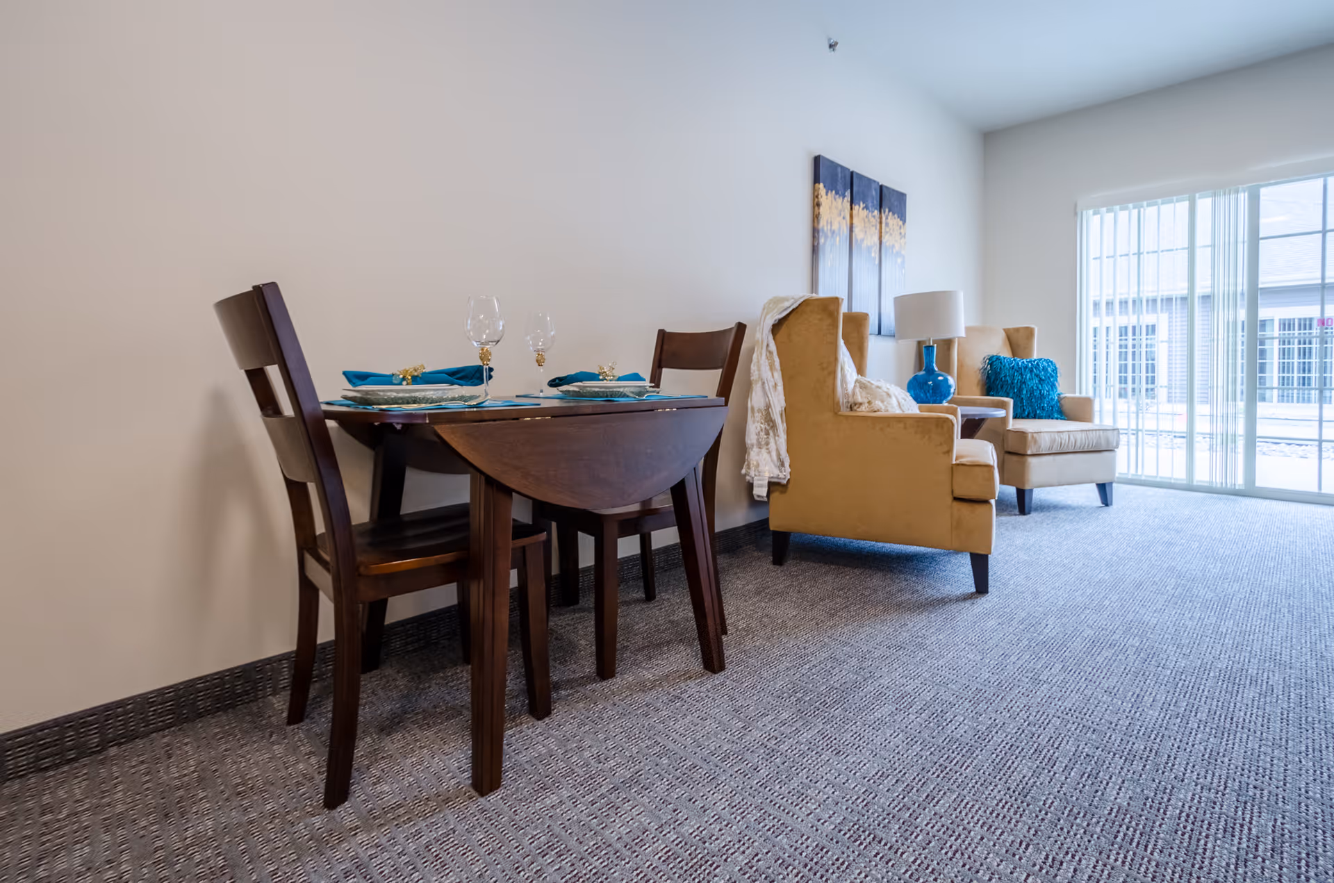 A small dining area with a dark wooden table set for two with blue napkins and wine glasses, adjacent to a living area with two beige armchairs, a blue lamp on a side table, and a large window with vertical blinds letting in natural light.