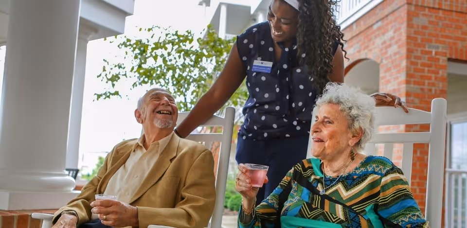 An elderly man and woman sitting on rocking chairs on a porch, each holding a drink, smiling and interacting with a standing caregiver who is smiling back at them.
