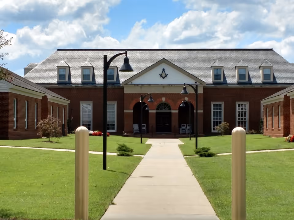 Front exterior view of a brick building with a gray shingled roof and multiple dormer windows. A paved walkway leads to the entrance, which has an archway and rocking chairs on the porch. There are black street lamps lining the walkway and green lawns on either side under a partly cloudy sky.