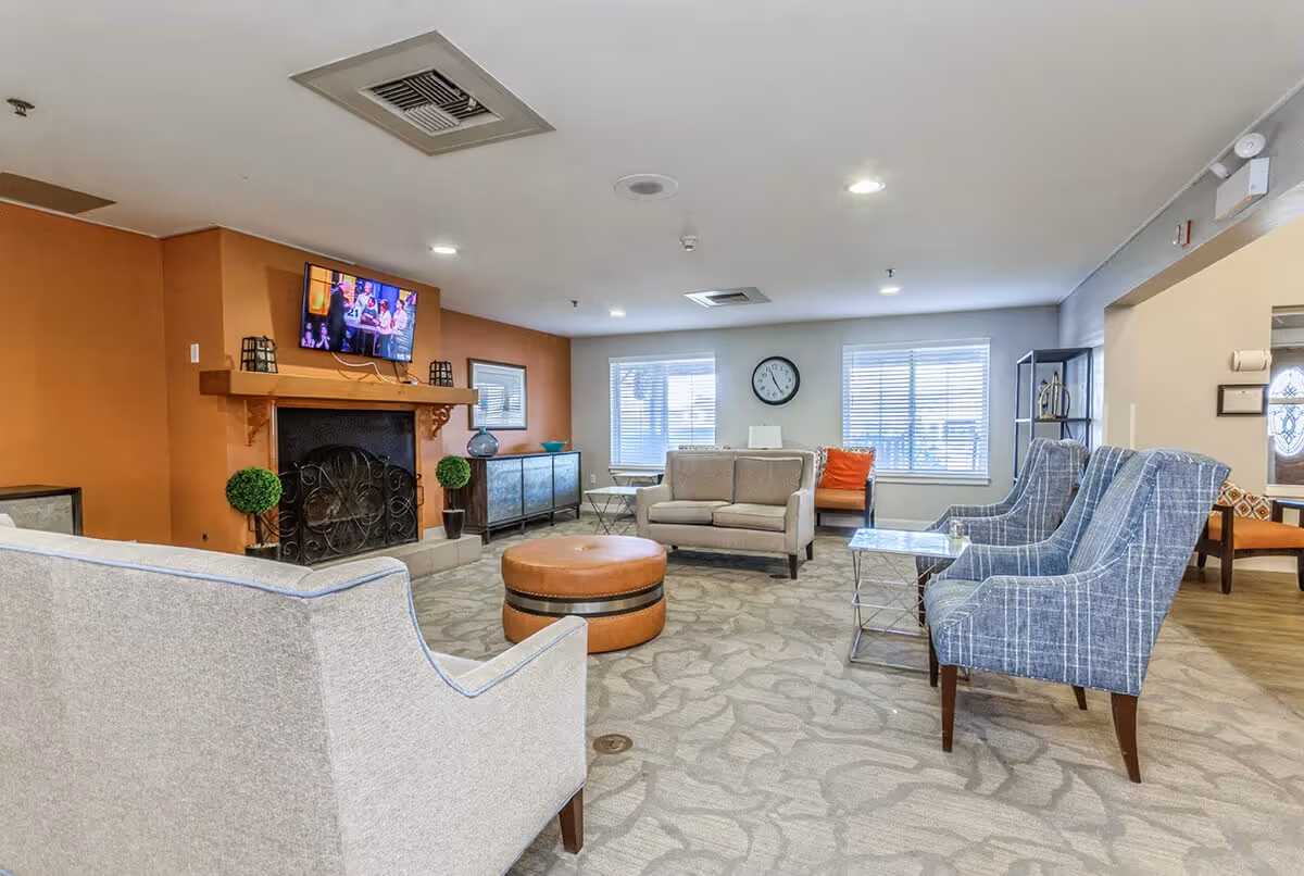 Communal lounge with sofas and armchairs arranged around a round ottoman, a fireplace with a TV above, and windows letting in daylight.
