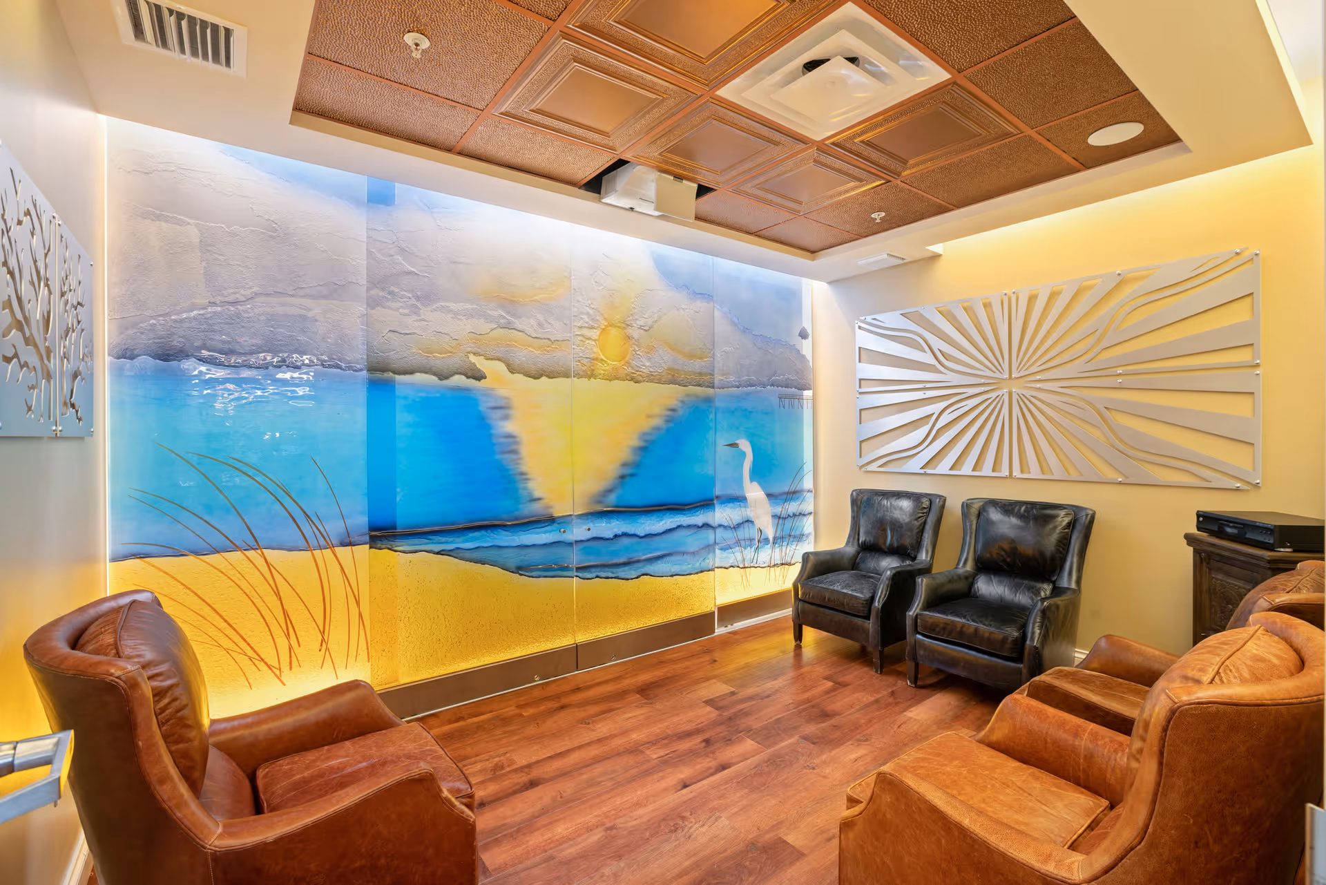Cozy seating room with leather armchairs facing a large colorful beach mural and decorative wall art.