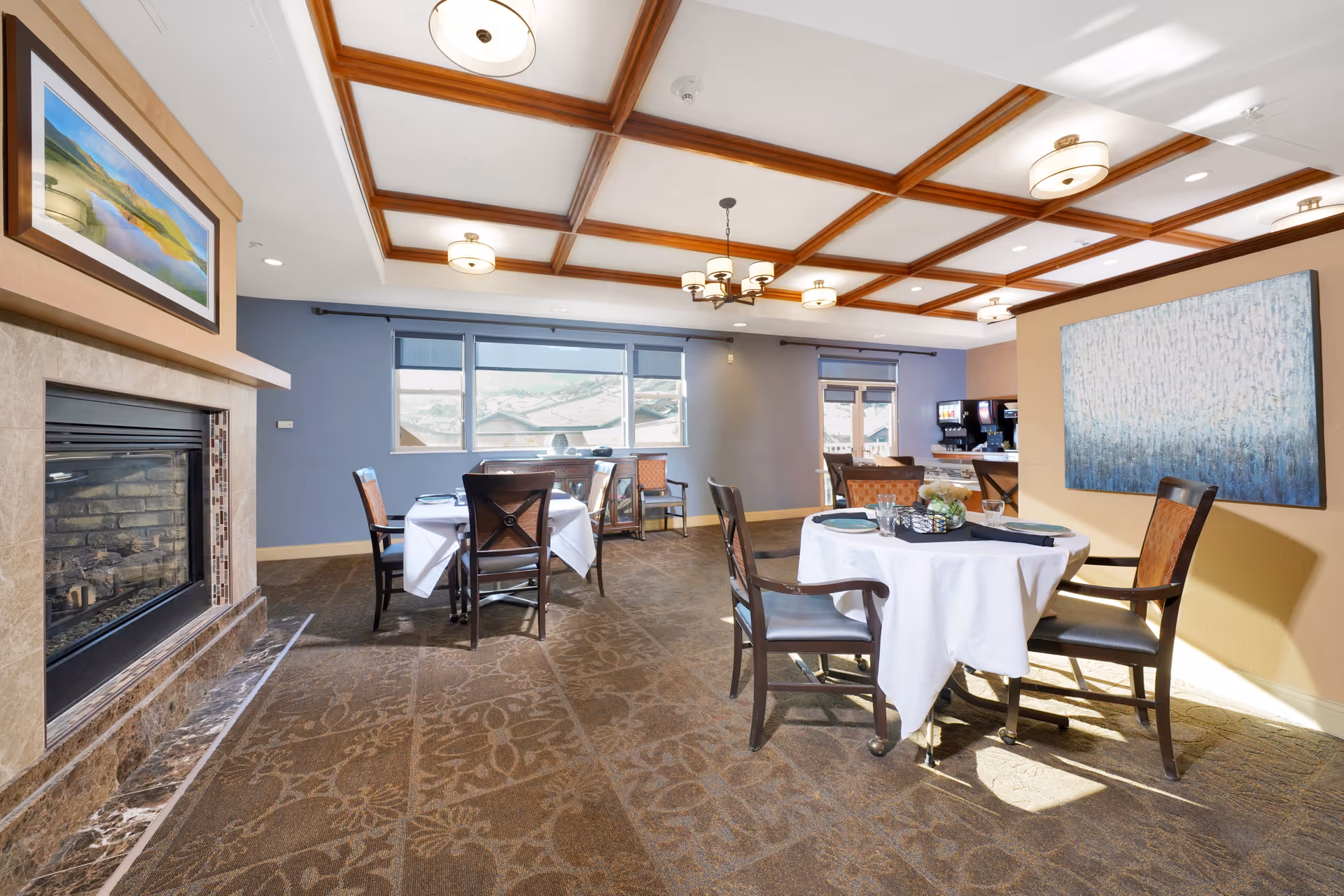 A bright dining room with round tables covered in white tablecloths, set with plates and glasses. The room features a coffered ceiling with multiple light fixtures, a fireplace with a landscape painting above it, large windows letting in natural light, and a beverage station in the background. The walls are painted in soft blue and beige tones, and there is a large abstract painting on one wall.