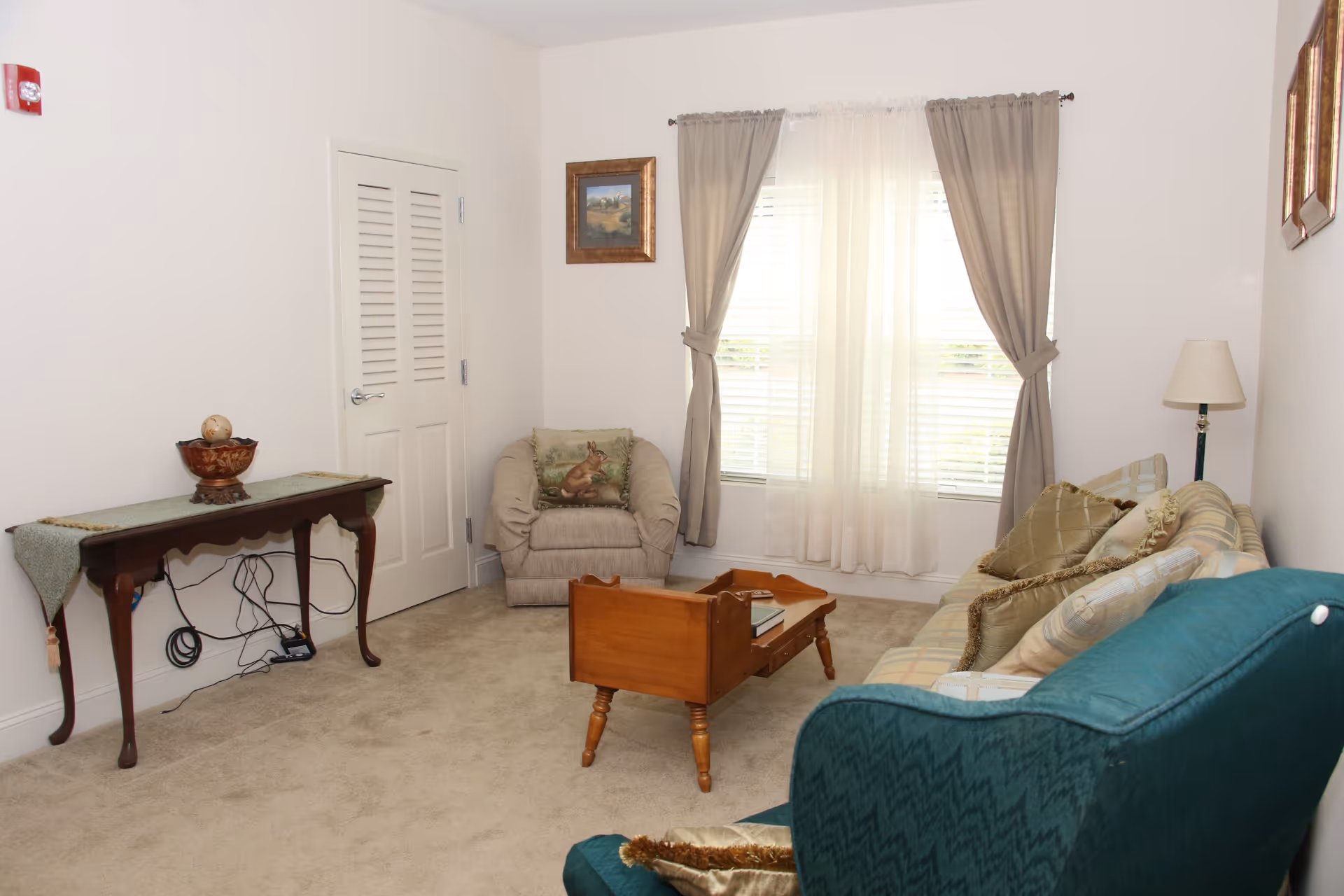 A cozy living room with beige carpet, a plaid sofa with multiple pillows, a green armchair, a wooden coffee table, a beige armchair with a decorative pillow, a wooden console table with a decorative bowl, a floor lamp, and a window with sheer white curtains and beige drapes.