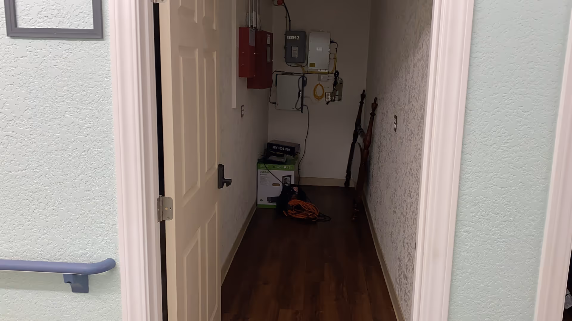 A narrow hallway with a partially open door on the left side. The hallway has wood flooring and textured white walls. At the end of the hallway, there are electrical panels mounted on the wall, a box, and some coiled cables on the floor.
