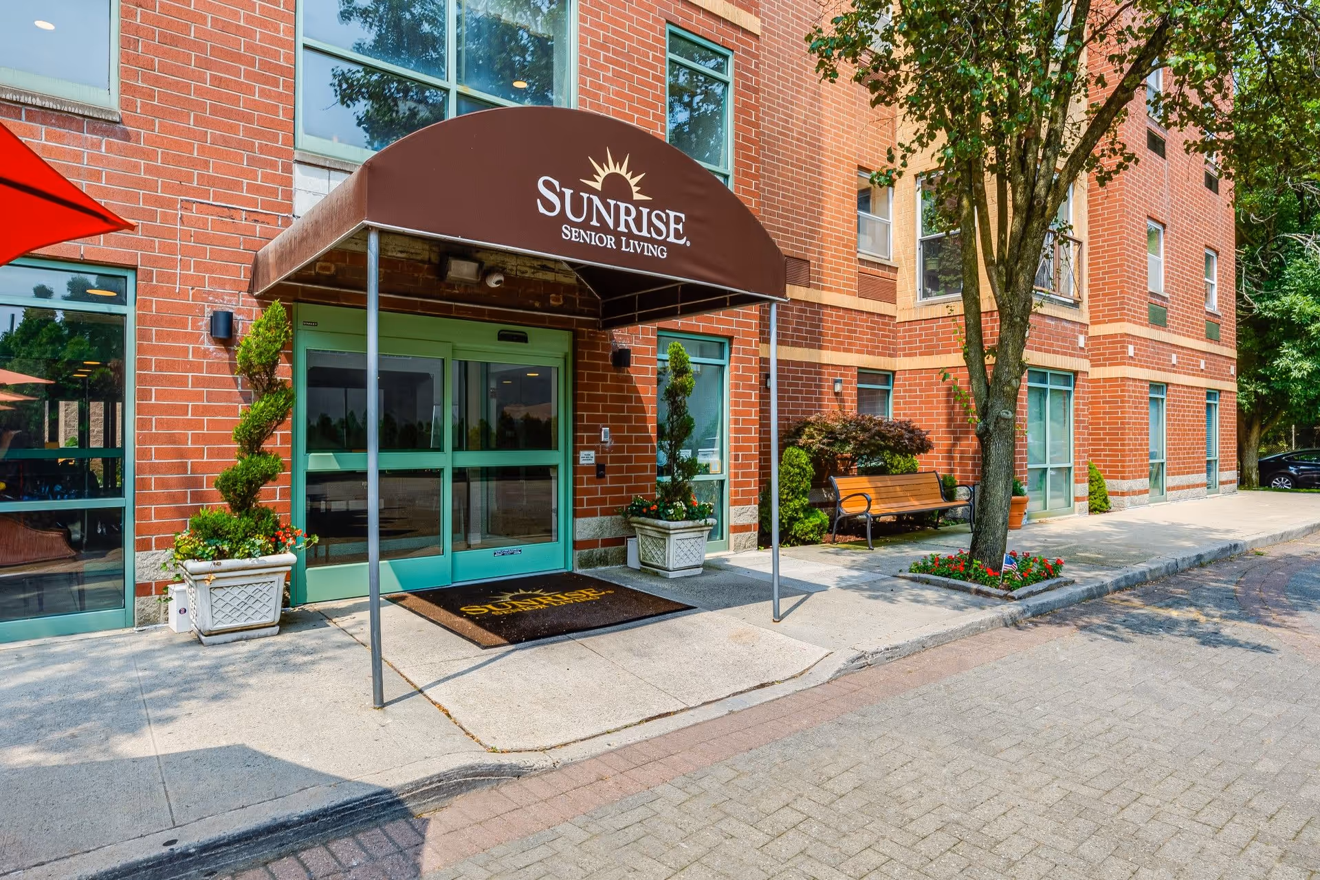 Front entrance of a brick Sunrise Senior Living building with a brown awning, green automatic doors, potted plants and a bench.