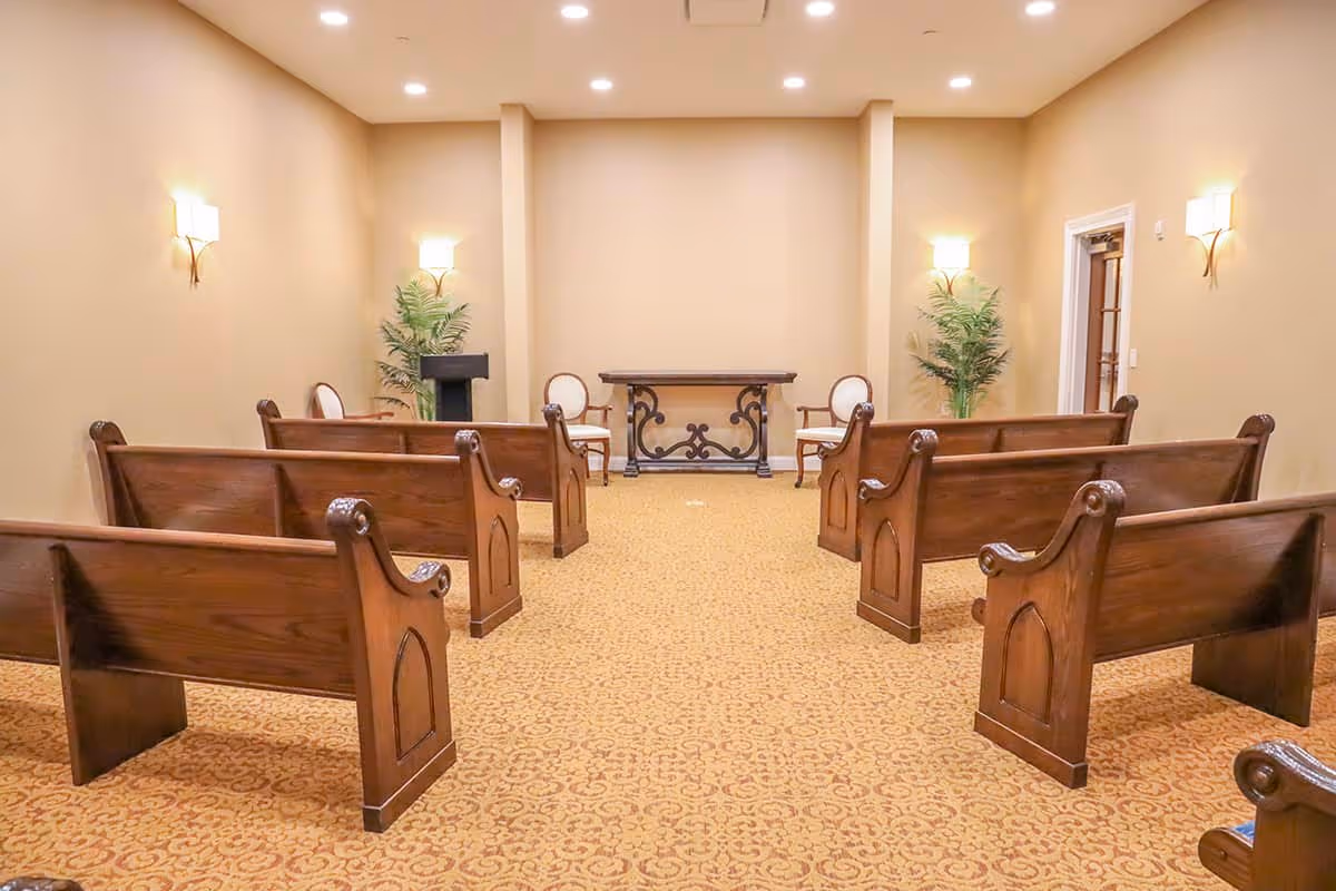 Interior view of a small chapel or meeting room with wooden pews arranged in rows facing a small table and two chairs at the front. The room has beige walls, patterned carpet, wall-mounted lights, and two potted plants near the front.