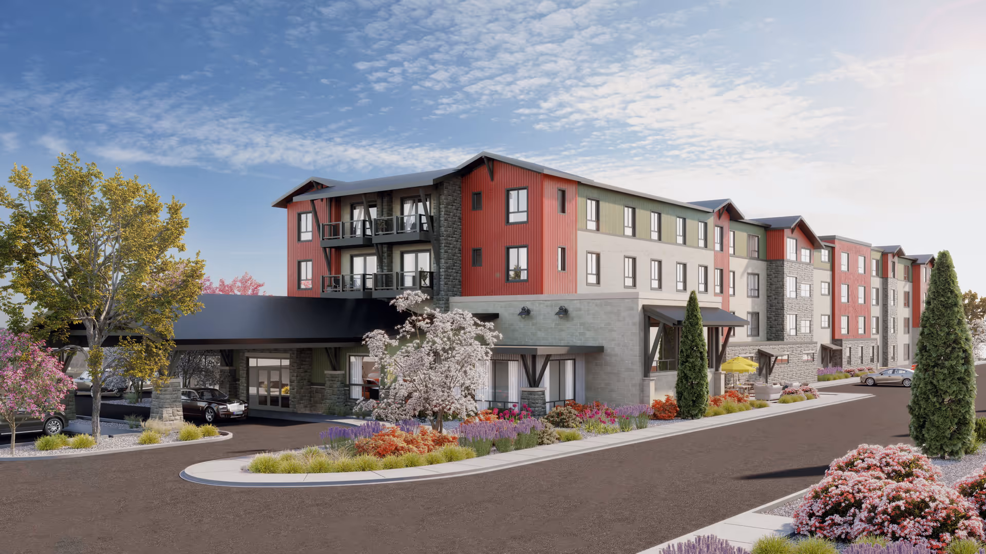 Exterior view of a modern senior living facility with a mix of red, green, and stone facade. The building has multiple floors with balconies and large windows. There is a covered entrance with cars parked underneath, surrounded by landscaped gardens with colorful flowers, trees, and shrubs. The sky is partly cloudy with bright sunlight.