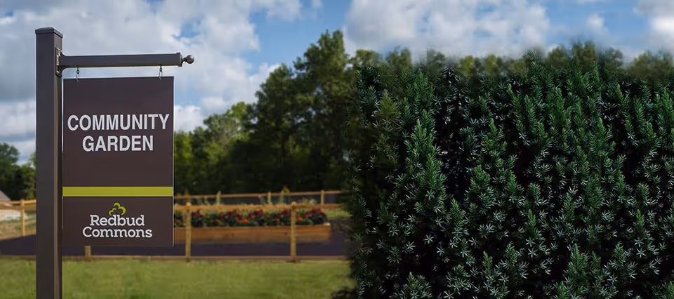 A signpost in a grassy outdoor area with a wooden fence and garden beds in the background. The sign reads 'COMMUNITY GARDEN' with the Redbud Commons logo below. The sky is partly cloudy and there are trees in the distance.