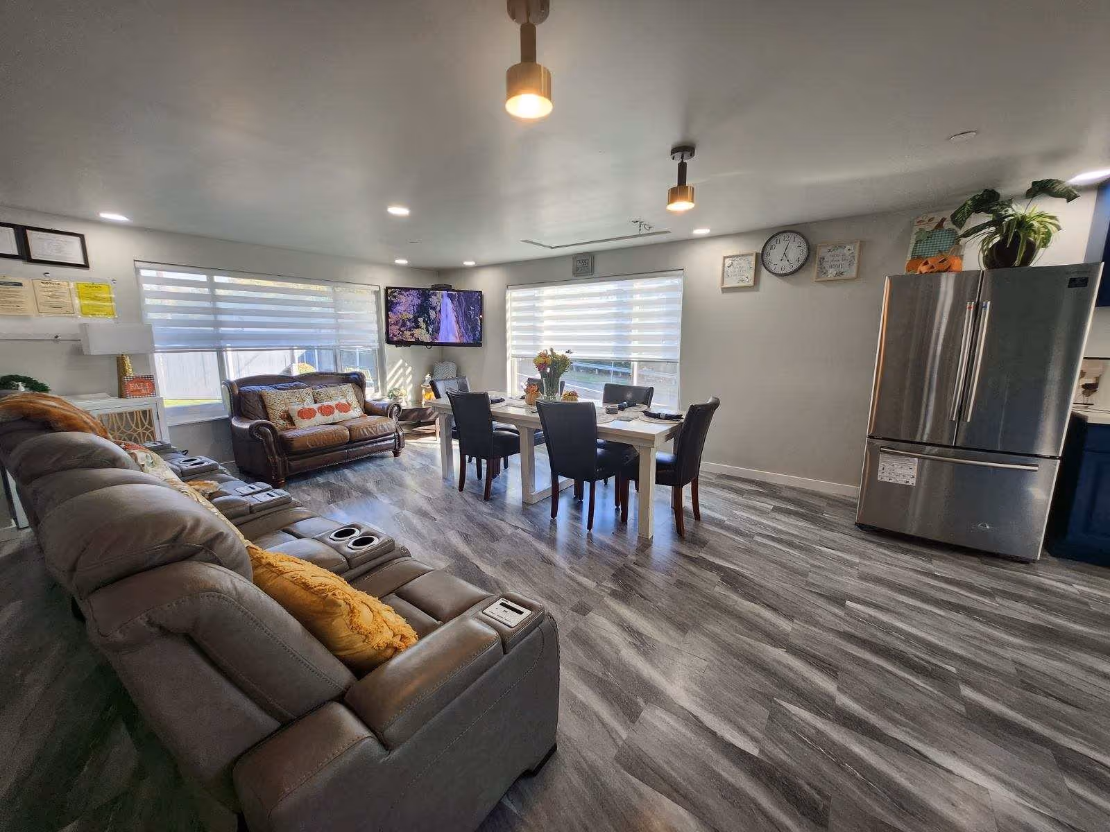A bright and spacious living and dining area with a gray reclining sofa and a brown leather loveseat. A white dining table with four black chairs is positioned near two large windows with blinds. A flat-screen TV is mounted on the wall between the windows. The room features gray wood-patterned flooring, a stainless steel refrigerator, and several decorative plants and wall hangings.