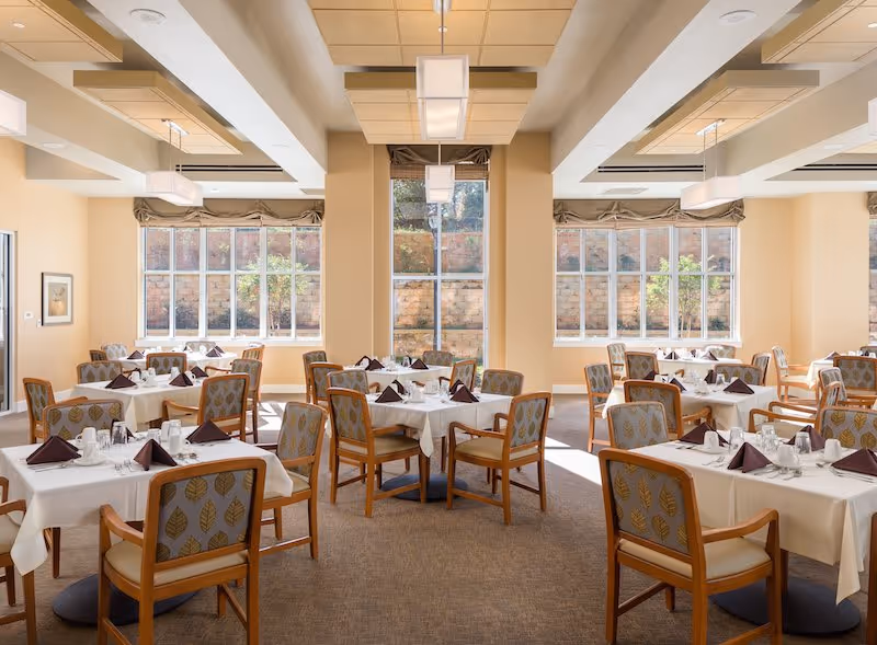 A bright dining room with multiple tables covered in white tablecloths, each set with folded dark napkins, glasses, and silverware. The room has large windows letting in natural light, beige walls, and patterned chairs with wooden frames.