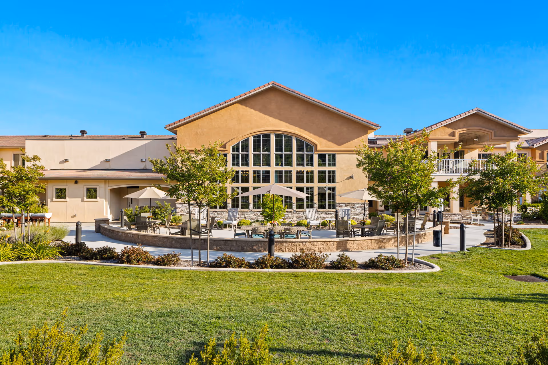 Outdoor patio area of a senior living facility with tables, chairs, and umbrellas surrounded by small trees and landscaping, in front of a building with large arched windows under a clear blue sky.