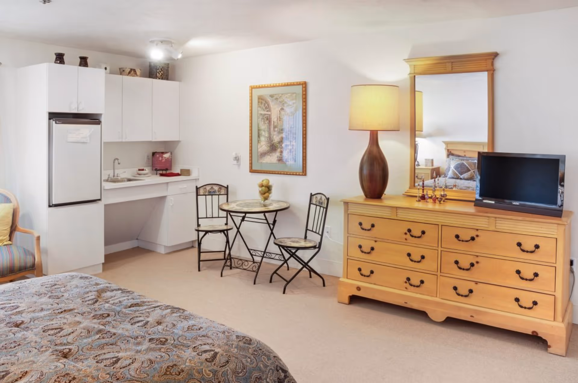 Interior view of a senior living facility room featuring a bed with patterned bedding in the foreground, a small kitchenette with white cabinets and a refrigerator, a round table with two chairs, and a wooden dresser with a large mirror and a lamp on top. A flat-screen TV is placed on the dresser, and a framed picture hangs on the wall.