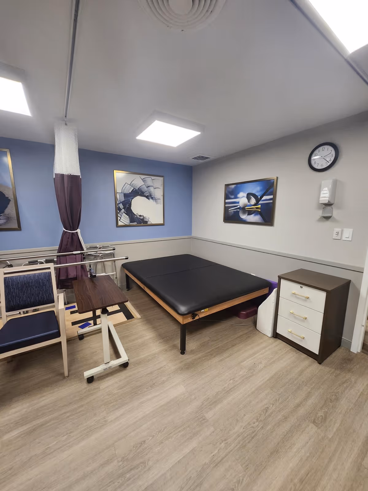 A room with a padded black therapy or examination table, a wooden chair with blue upholstery, a small adjustable table on wheels, a three-drawer cabinet, and two framed abstract pictures on the walls. The room has light wood flooring, a clock on the wall, and a hand sanitizer dispenser.