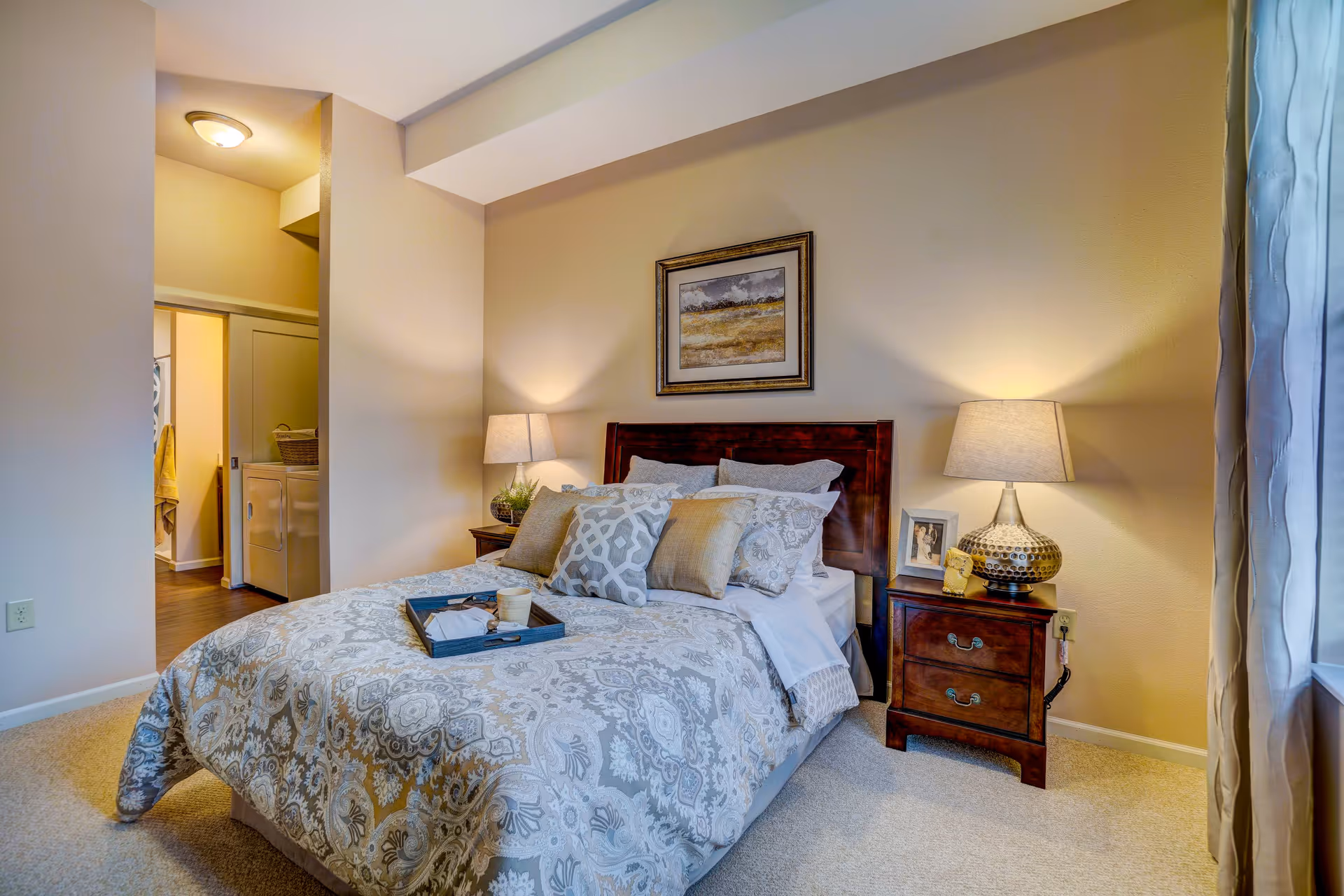 A cozy bedroom with a neatly made bed featuring patterned bedding and multiple pillows. There are two wooden nightstands on either side of the bed, each with a lamp. A framed picture hangs above the bed. To the left, there is an open doorway leading to another room with a washing machine and dryer visible. Light beige walls and carpeted floor complete the room.
