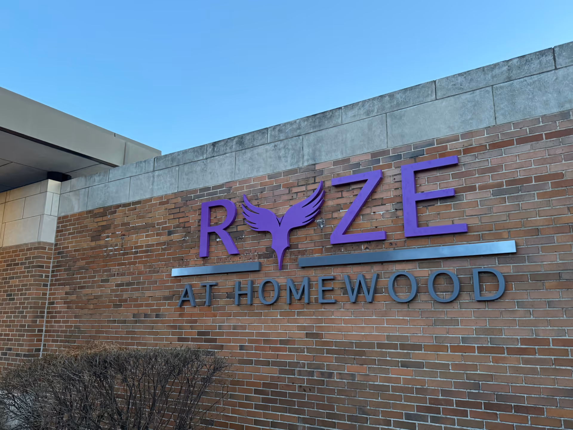 Exterior brick wall with a purple RYZE AT HOMEWOOD sign and winged logo.