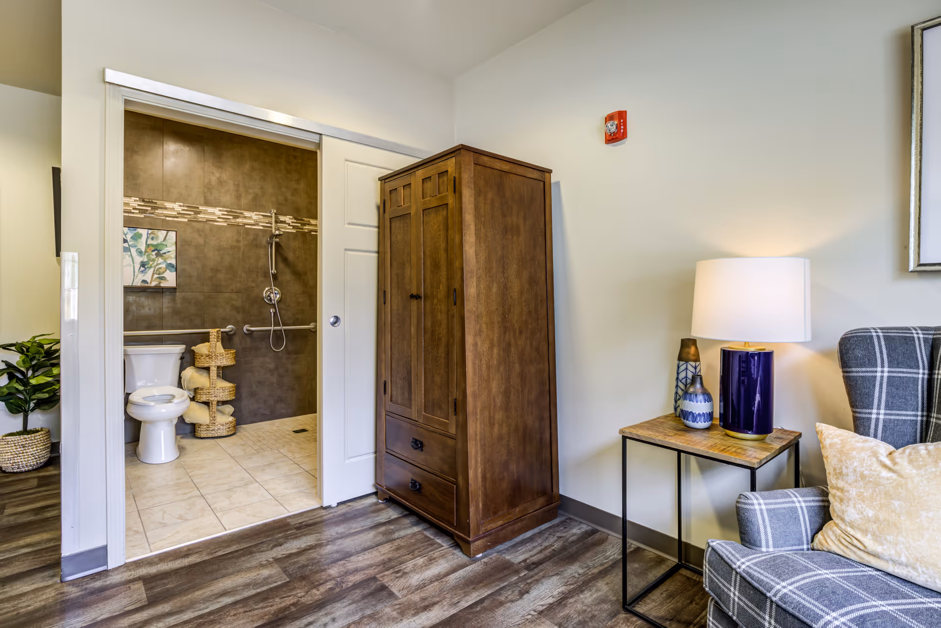 View of a cozy room with a wooden wardrobe, a side table with a lamp and decorative vases, and a plaid armchair with a pillow. Through an open sliding door, a bathroom is visible with a toilet, a walk-in shower with grab bars, and a piece of wall art.