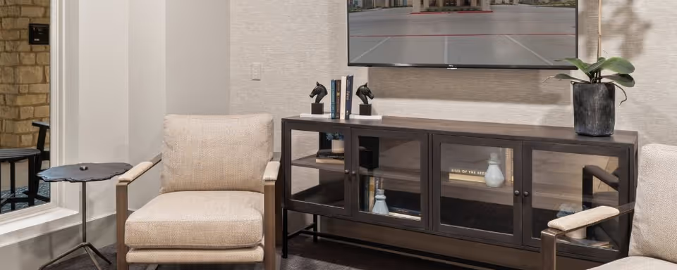 Cozy living room area with beige armchairs, a dark media console under a wall-mounted TV, and a small side table with a potted plant.