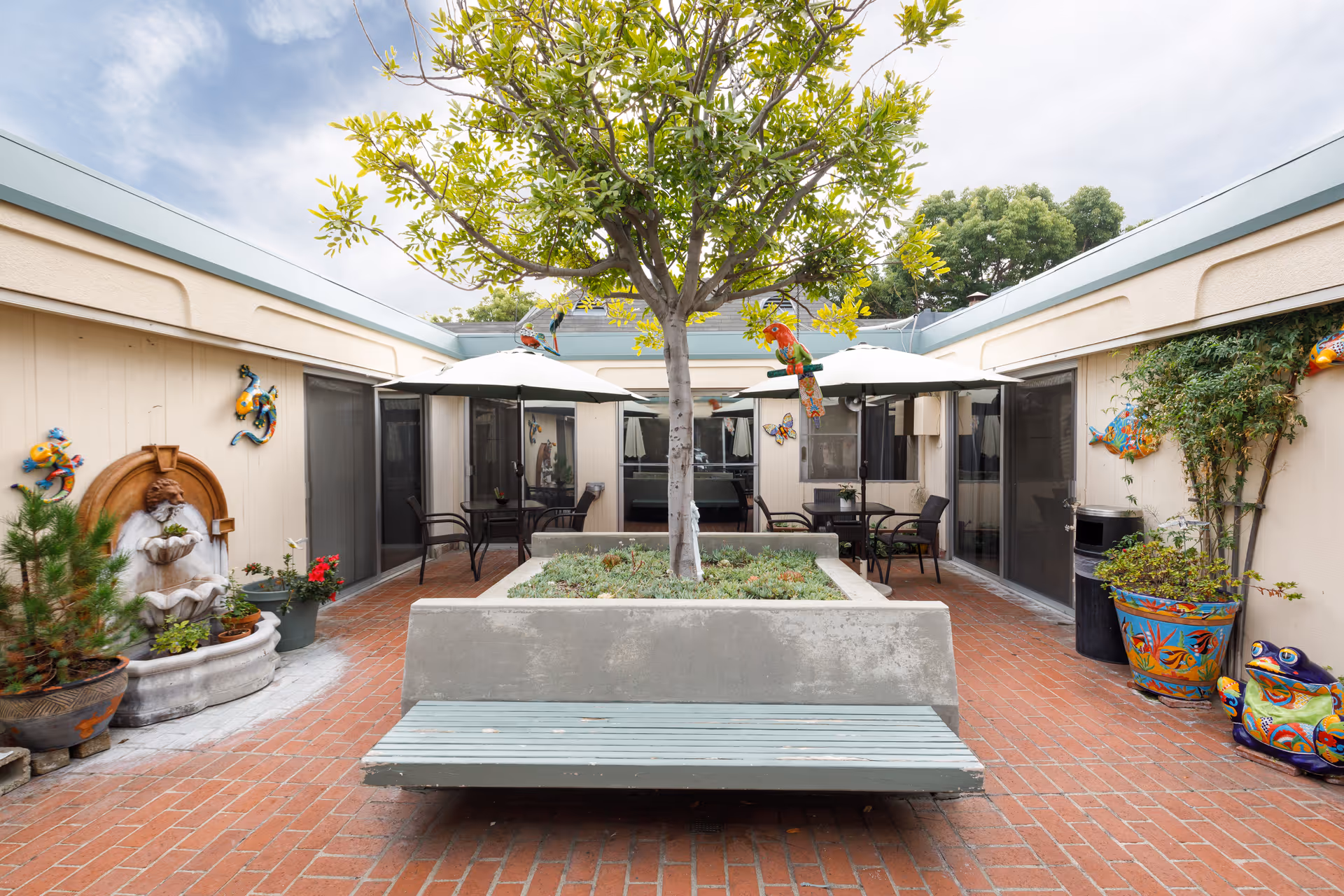 Outdoor courtyard area with a central tree planted in a raised concrete planter that has a bench attached. The courtyard is surrounded by beige walls with sliding glass doors and decorated with colorful ceramic animal wall art and potted plants. There are two patio tables with umbrellas and chairs on either side of the courtyard.