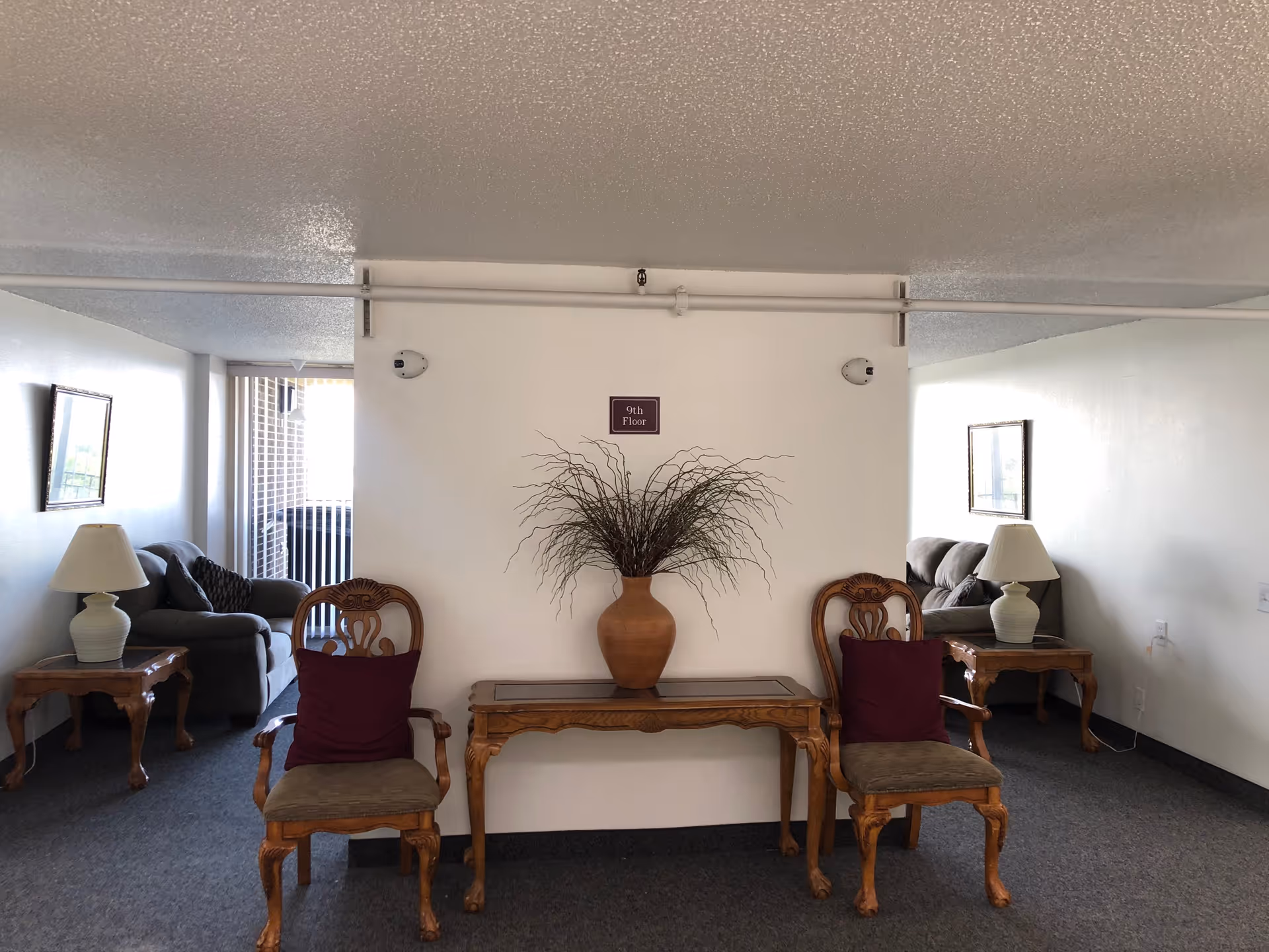 A hallway area on the 9th floor with two wooden chairs with purple cushions, a wooden table with a large vase containing decorative branches, two side tables with lamps, and two couches in alcoves on either side.