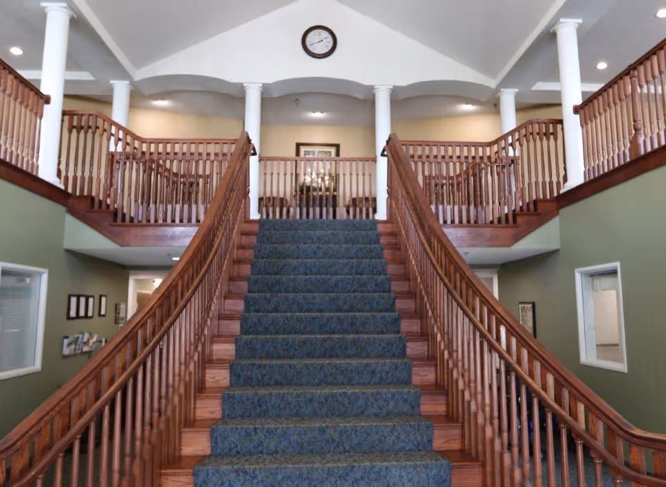 A grand interior staircase with blue carpet and wooden railings leading up to a second-floor balcony with white columns and a clock on the wall.