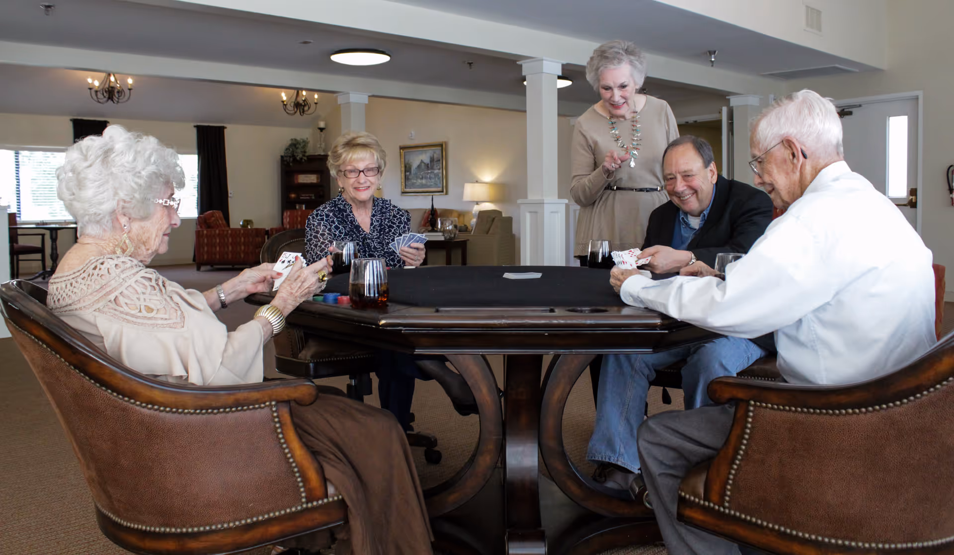 A group of four elderly people sitting around a card table playing cards, with one elderly woman standing and smiling. The room is well-lit with comfortable chairs and homey decor, including lamps, paintings, and curtains.