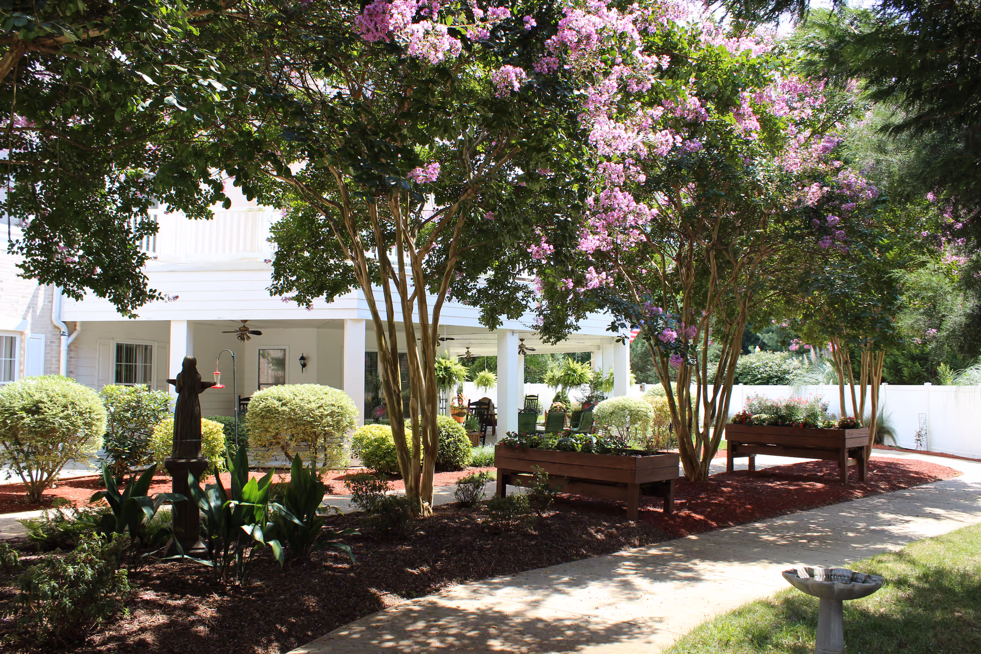 A peaceful outdoor garden area at HeartFields at Cary with blooming pink flowers on trees, neatly trimmed bushes, raised wooden flower beds, a small statue, and a birdbath. A white building with a covered porch and ceiling fans is visible in the background.