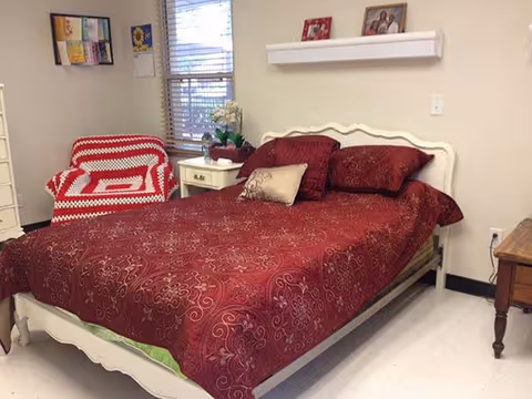 A bedroom with a white wooden bed frame and a red patterned bedspread with matching pillows. Next to the bed is a white nightstand with a plant and a small lamp. There is a red and white crocheted blanket draped over a chair near a window with blinds. The walls are beige with a small shelf holding framed pictures.