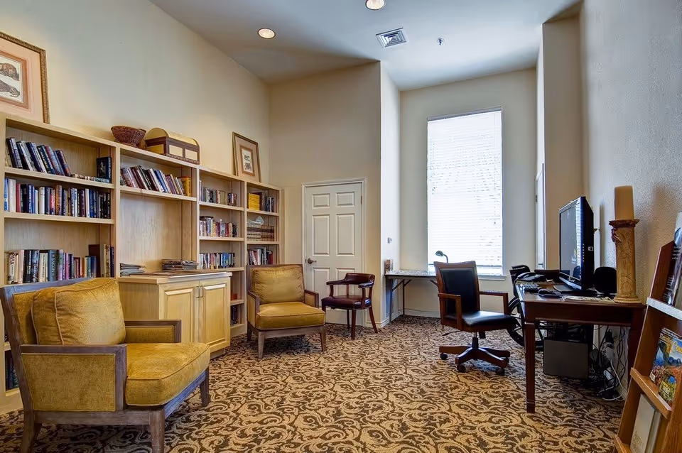 Bright communal library and computer room with bookshelves, armchairs, patterned carpet, and desks with computers.