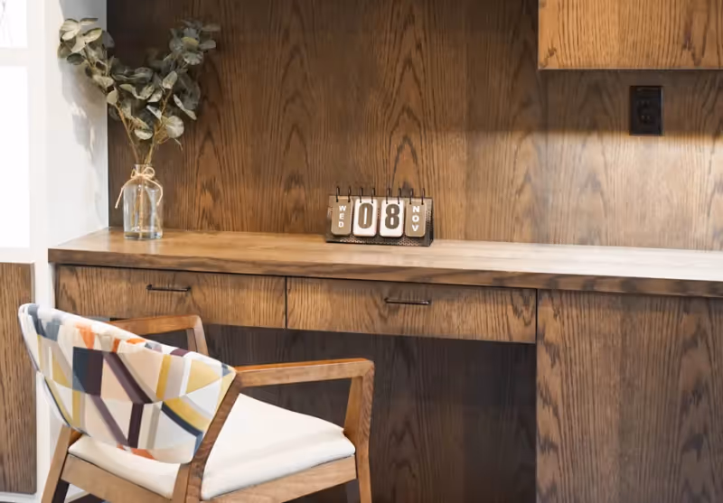 A wooden desk with drawers and a matching wooden wall panel behind it. On the desk is a small glass vase with dried eucalyptus branches and a flip calendar showing Wednesday, November 8. A wooden chair with a white cushion and a colorful geometric patterned backrest is positioned in front of the desk.