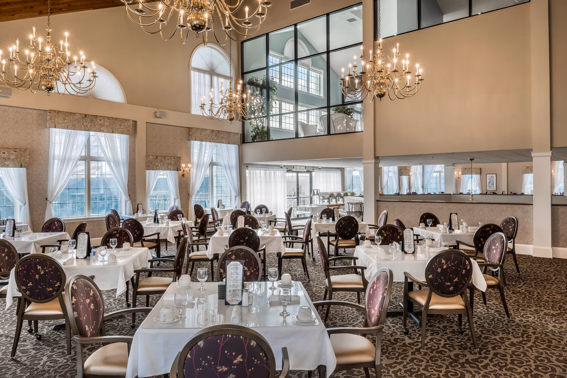 Elegant dining room with multiple tables covered in white tablecloths, set with glassware, cups, and menus. The room features large windows with sheer curtains, ornate chandeliers hanging from a high ceiling, and patterned carpet flooring. Chairs have dark, floral-patterned upholstery.
