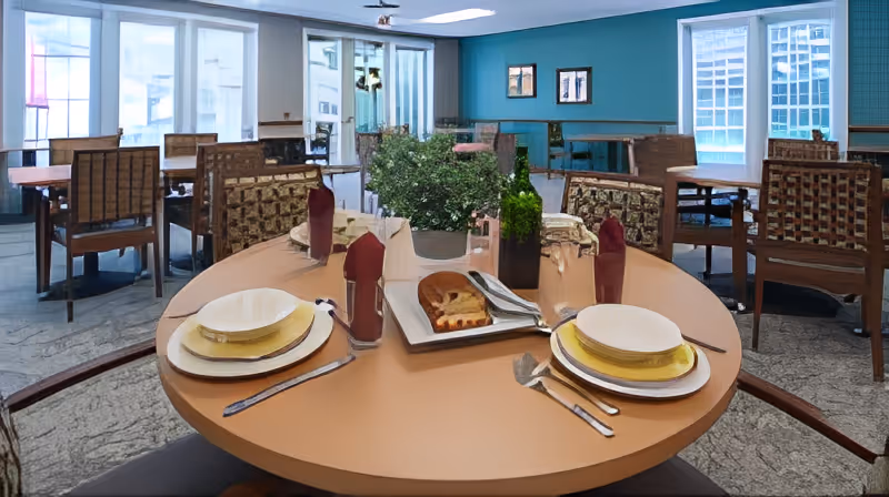 A round dining table set with plates, cutlery, napkins, and a plant centerpiece in a bright communal dining room with multiple tables and large windows.