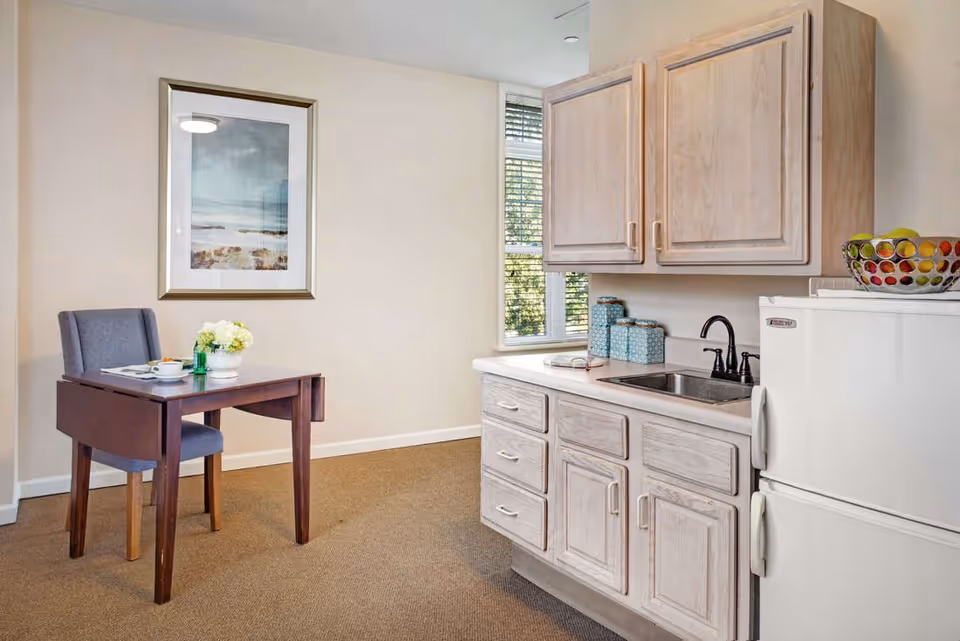 A small kitchen area with light wood cabinets, a white refrigerator, and a sink with a black faucet. Next to the kitchen is a wooden table with a gray upholstered chair, a cup, a small vase with flowers, and a framed picture on the wall. A window with blinds lets in natural light.