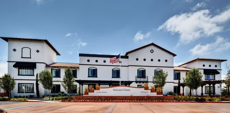 Front exterior view of a large, white senior living facility building with a red-tiled roof, multiple windows, and an American flag flying above the entrance. There are trees and landscaping in front of the building and a sign that reads 'Welcome Home' with directions to the main entrance and parking.