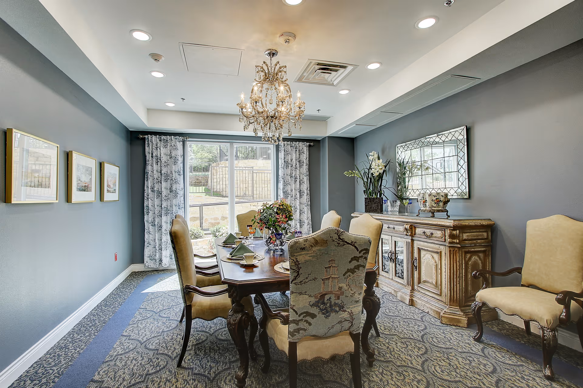Well-appointed dining room with a wooden table and upholstered chairs under a chandelier, a sideboard, and a large window with curtains.