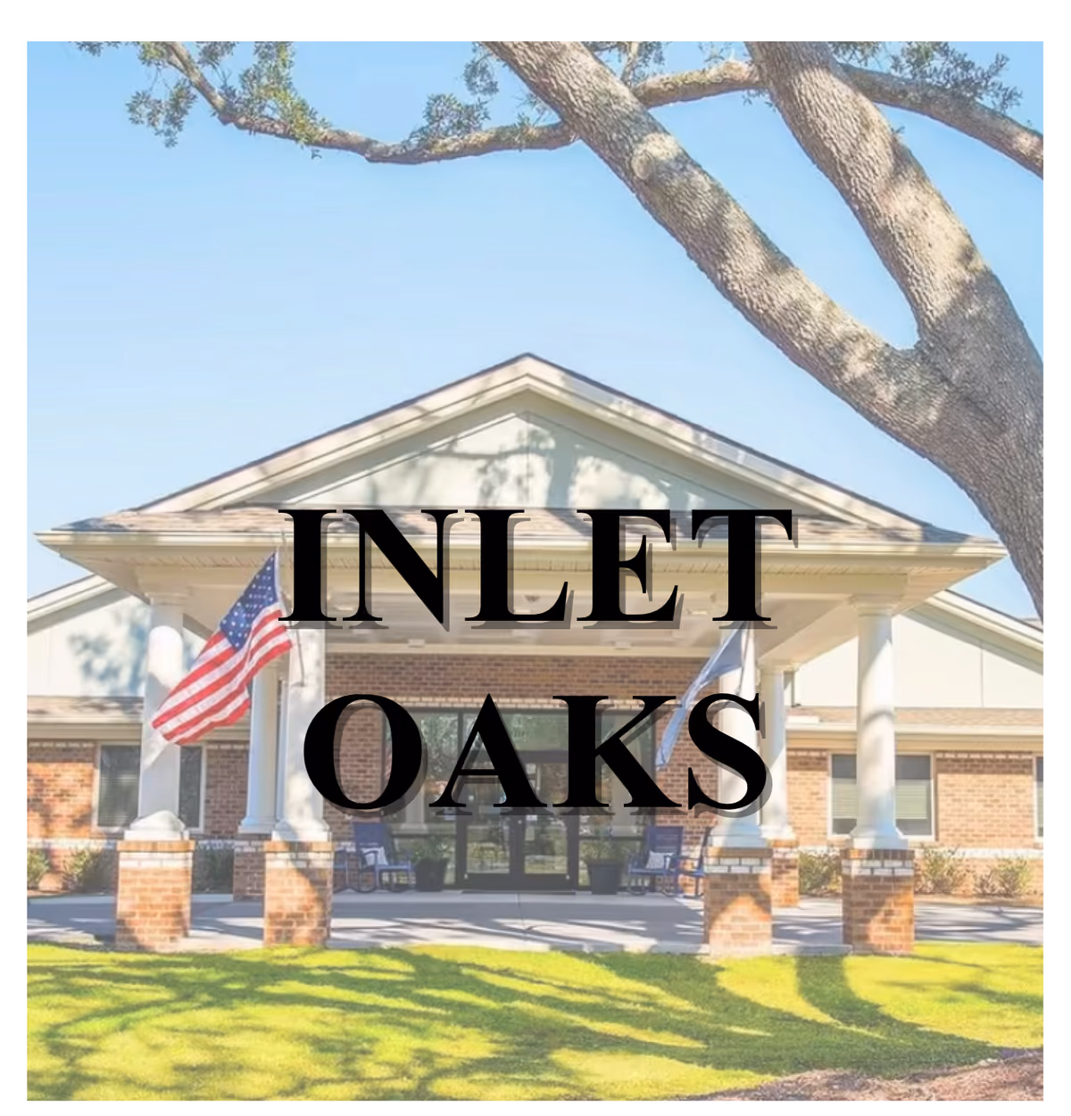 Front exterior view of a single-story brick building with white columns supporting a covered entrance. Two flags, including an American flag, are displayed near the entrance. A large tree branch extends over the building, and there is green grass in the foreground.