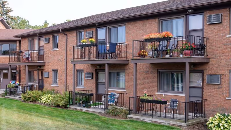 Exterior view of a two-story brick senior living facility with balconies and patios decorated with potted plants and outdoor furniture, surrounded by green grass and shrubs.