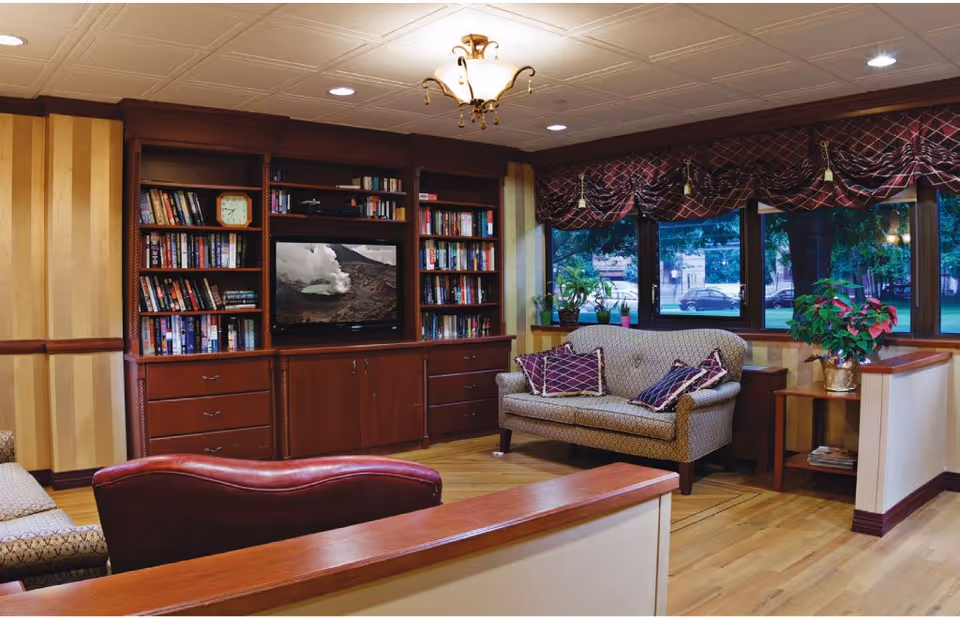 A cozy living room area with wooden bookshelves filled with books and a TV displaying a nature scene. There is a patterned loveseat with decorative pillows, a side table with a potted plant, and large windows with valance curtains showing an outdoor view with trees and parked cars.
