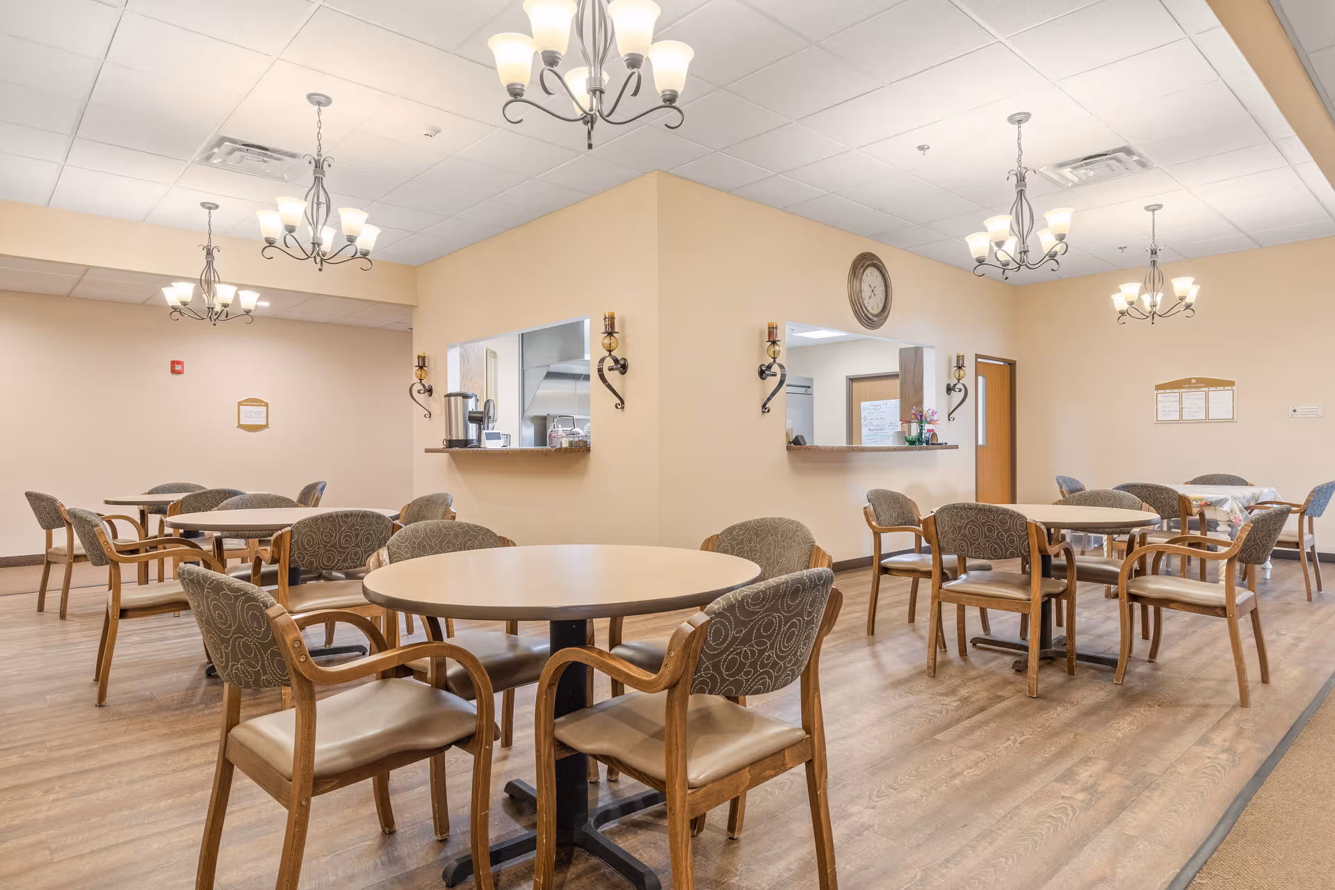 Dining room with round tables and chairs, chandeliers, wood flooring, and a serving pass-through window.