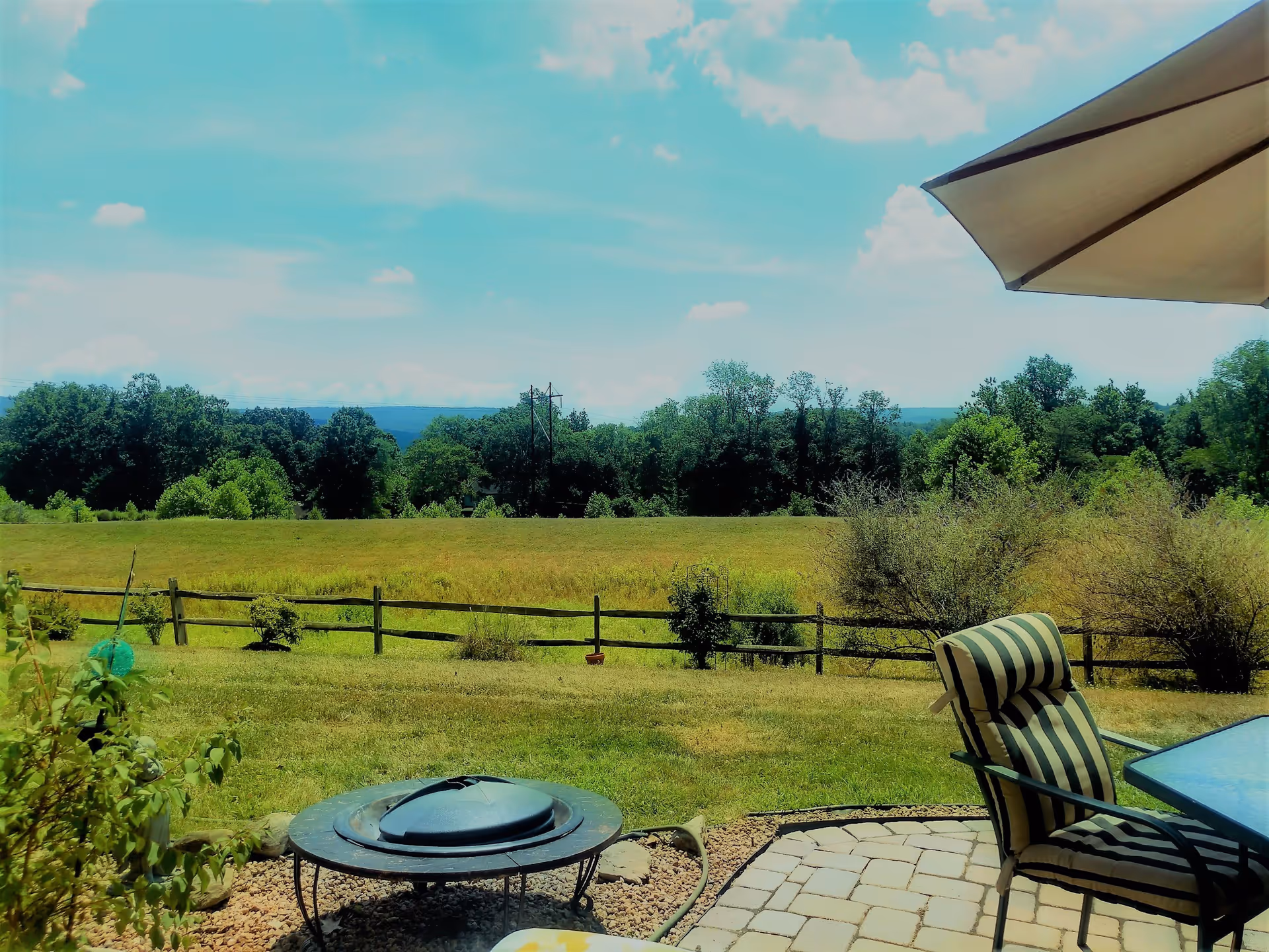 Outdoor patio area with a striped cushioned chair and glass table under a large umbrella, overlooking a grassy field with a wooden fence and trees in the background under a partly cloudy sky.