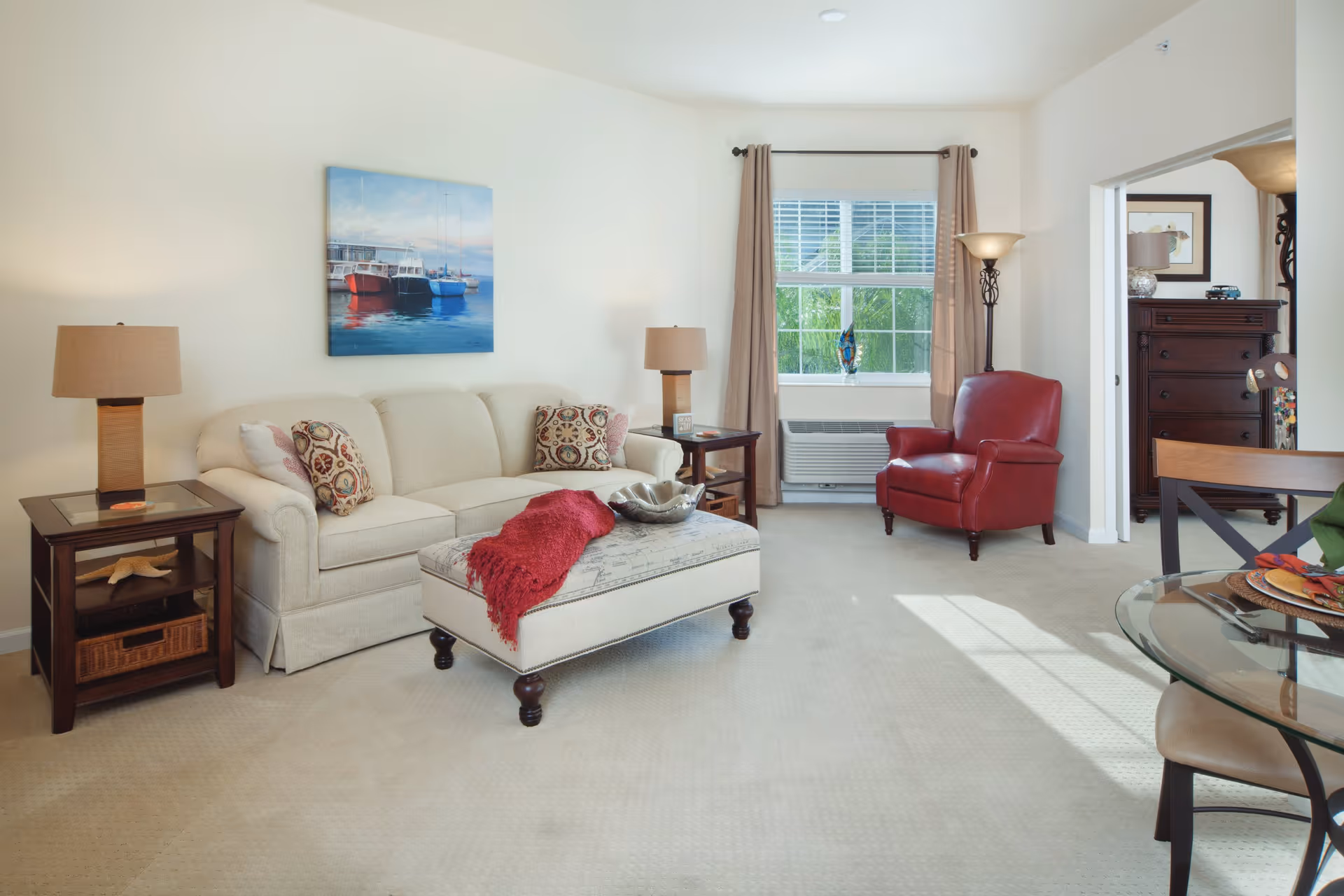 A bright and cozy living room with a beige sofa adorned with patterned cushions, a red throw blanket on a cushioned ottoman, a red leather armchair near a window with beige curtains, two wooden side tables with lamps, a floor lamp, and a glass-top dining table with chairs partially visible. A painting of boats on water hangs on the wall above the sofa.