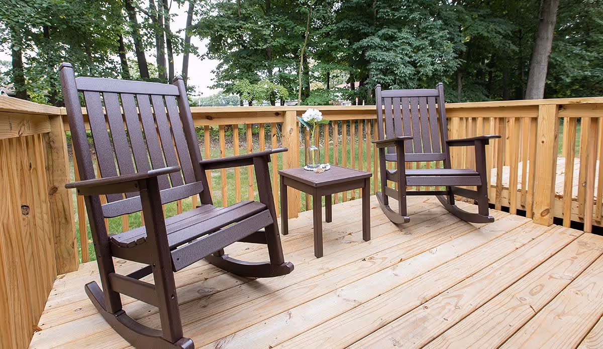 Two brown wooden rocking chairs on a wooden deck with a small matching table between them holding a glass vase with white flowers. The deck is surrounded by a wooden railing and trees are visible in the background.