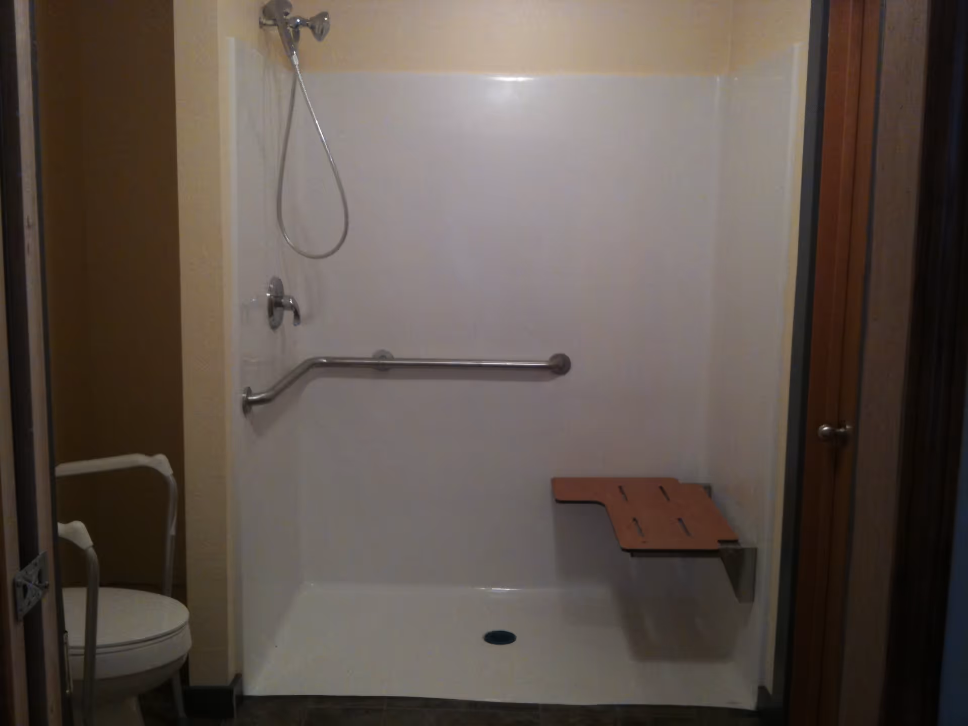 Accessible shower area with a handheld showerhead, a horizontal grab bar, and a fold-down wooden shower seat. To the left, a toilet with a white safety frame is partially visible.