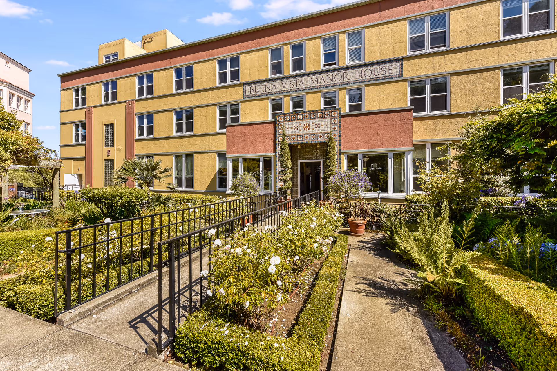 Exterior view of Buena Vista Manor House, a three-story yellow and red building with multiple windows. The entrance is decorated with colorful tiles and flanked by two tall, trimmed bushes. A paved walkway with black metal railings leads through a well-maintained garden with green hedges, white flowers, and various plants under a clear blue sky.