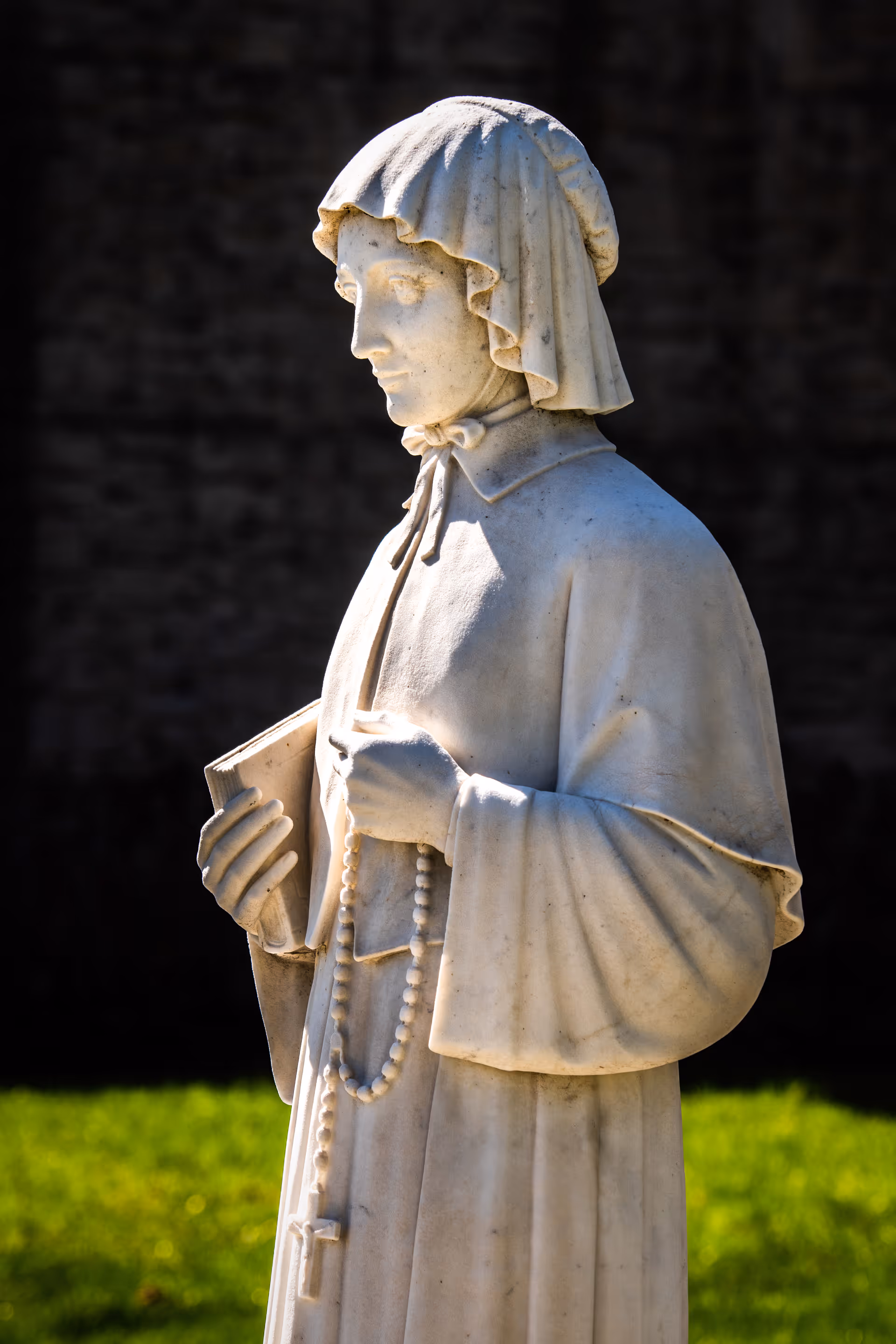 A white stone statue of a person wearing a hooded cloak, holding a book in one hand and a rosary with a cross in the other, standing outdoors on green grass with a dark blurred background.