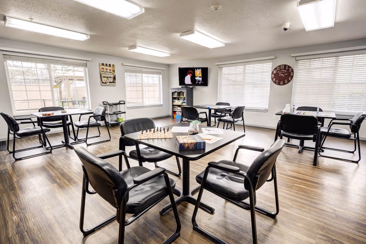 A bright activity room with multiple tables and black chairs arranged around them. On the closest table, there is a chessboard, a book, and some papers. The room has large windows with white blinds letting in natural light, a wall clock, and a TV mounted on the wall showing a person painting. The floor is wooden, and the room appears clean and organized.