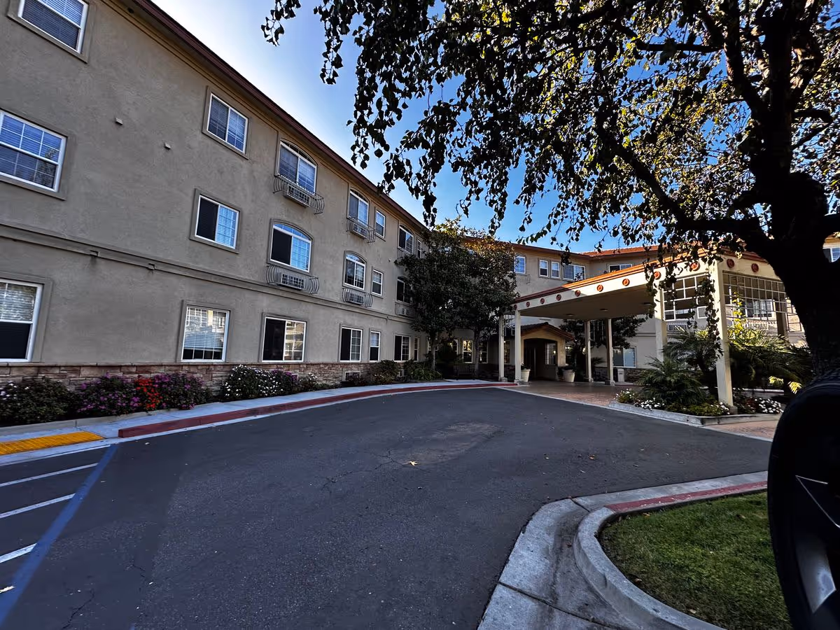 Front driveway and covered entrance of a three-story beige senior living building framed by trees.