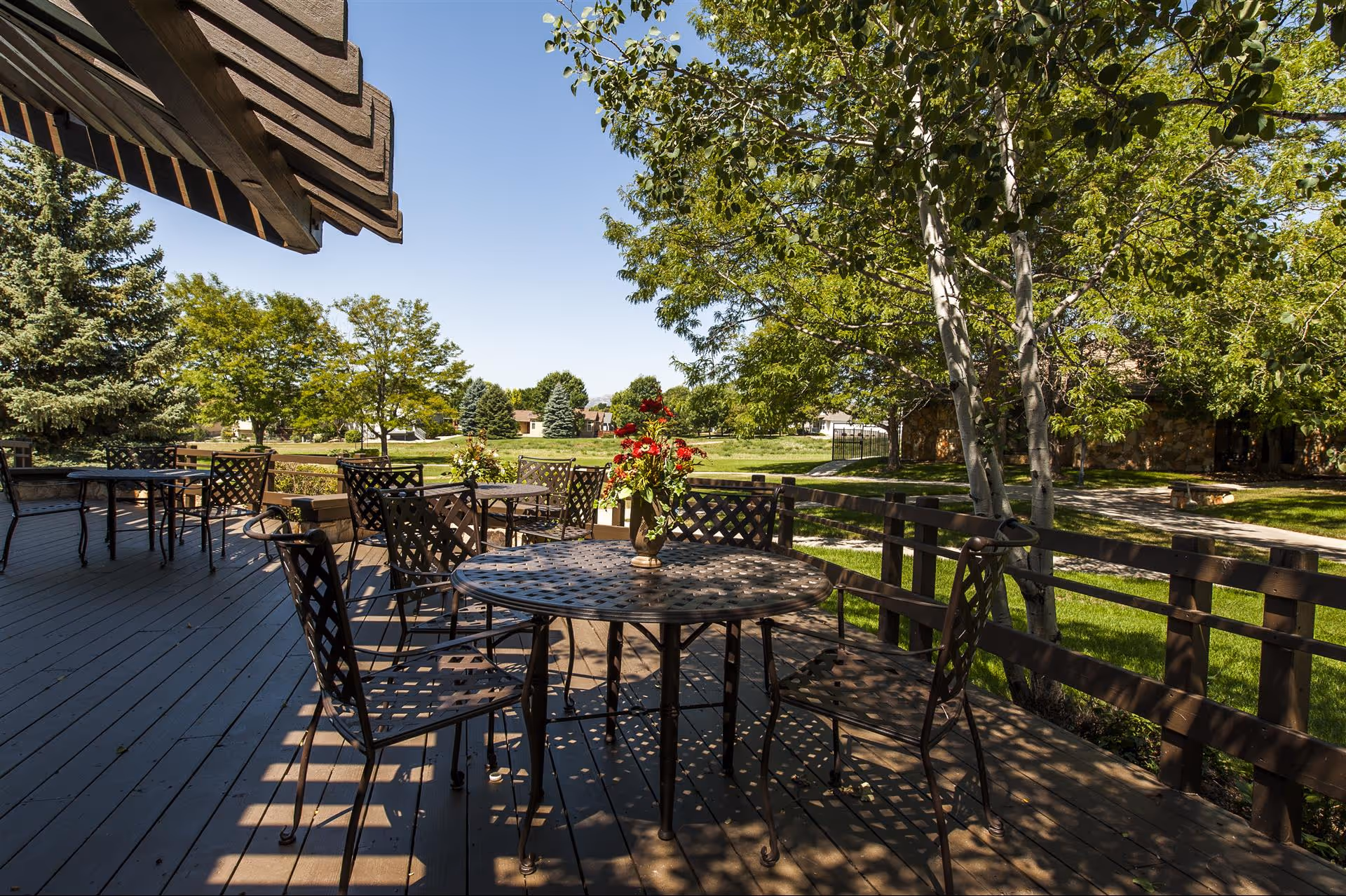 Outdoor patio area with metal tables and chairs on a wooden deck, surrounded by green trees and grass under a clear blue sky. A vase with red flowers is placed on one of the tables.