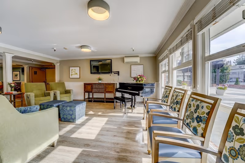 A bright and spacious living room area with large windows letting in natural light. The room features a black grand piano with a bouquet of flowers on top, a wooden sideboard, a wall-mounted TV, and several chairs including green armchairs, blue patterned ottomans, and wooden chairs with floral upholstery. The floor is light wood, and the walls are painted beige with white trim.