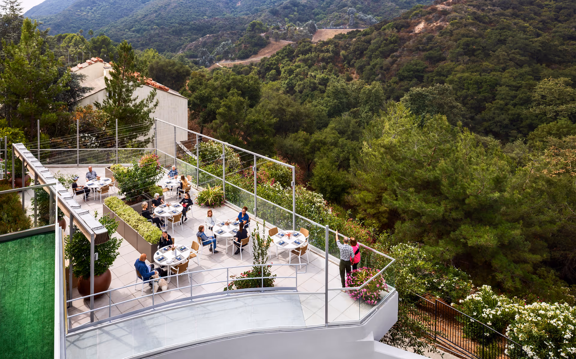 Outdoor terrace at Ciela Senior Living with several round tables and chairs where elderly people are sitting and socializing. The terrace is surrounded by greenery and overlooks a forested hillside with mountains in the background.