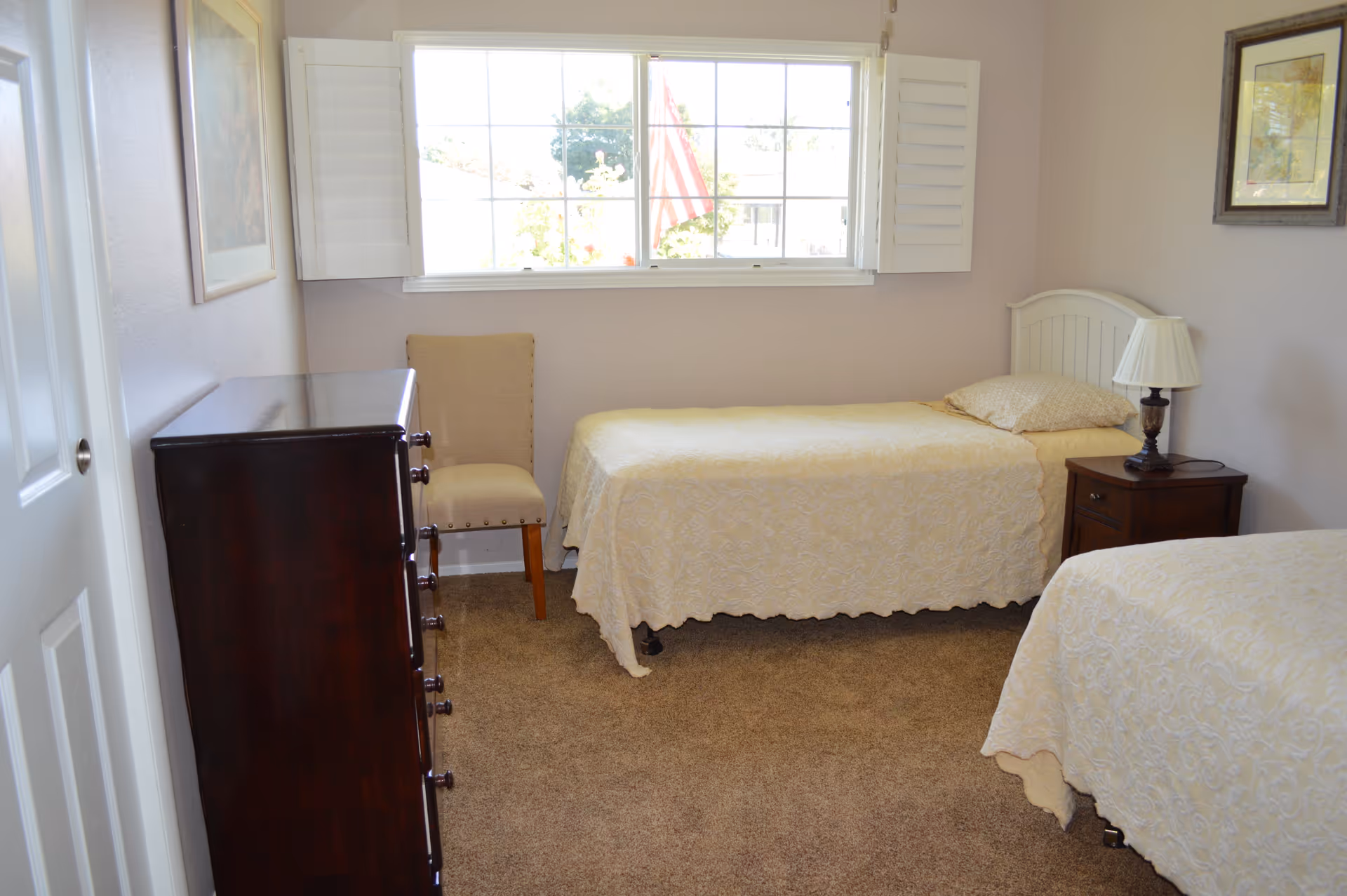 A small bedroom with two single beds covered in cream-colored bedspreads. There is a wooden nightstand with a lamp between the beds, a wooden dresser on the left side, and a beige upholstered chair near the window. The window has white shutters and an American flag is visible outside. The walls are light-colored and there are framed pictures hanging on them.